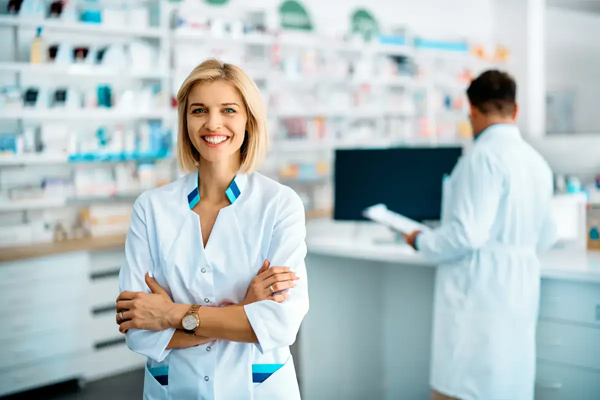 Farmacista sorridente in camice bianco in piedi in una farmacia, con un collega al bancone sullo sfondo